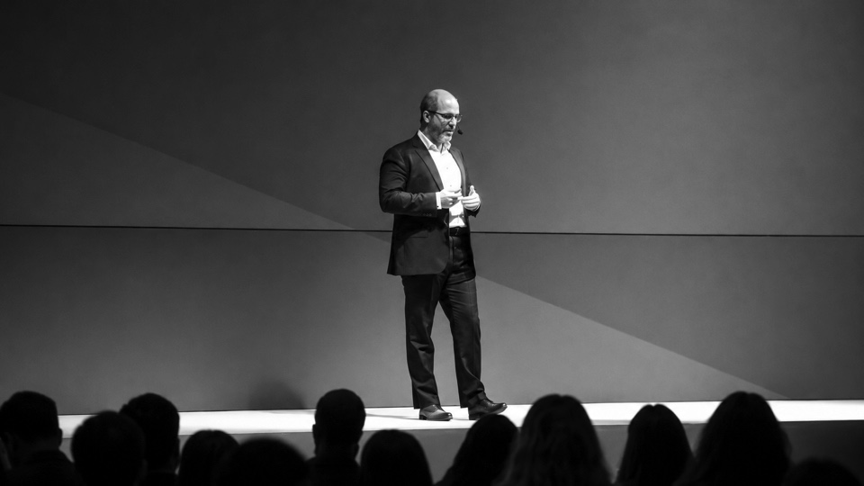 Sébastien Bourguignon seul sur scène lors d’une conférence, photographie noir et blanc, public en silhouette au premier plan.